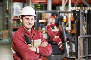 young smiling warehouse worker driver in uniform in front of forklift stacker loader